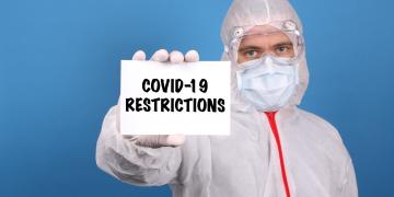 Medical doctor holding banner with Covid-19 Restrictions text, Isolated over blue background