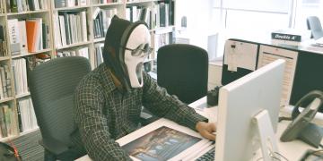 Masked man sitting behind a desk, using a computer